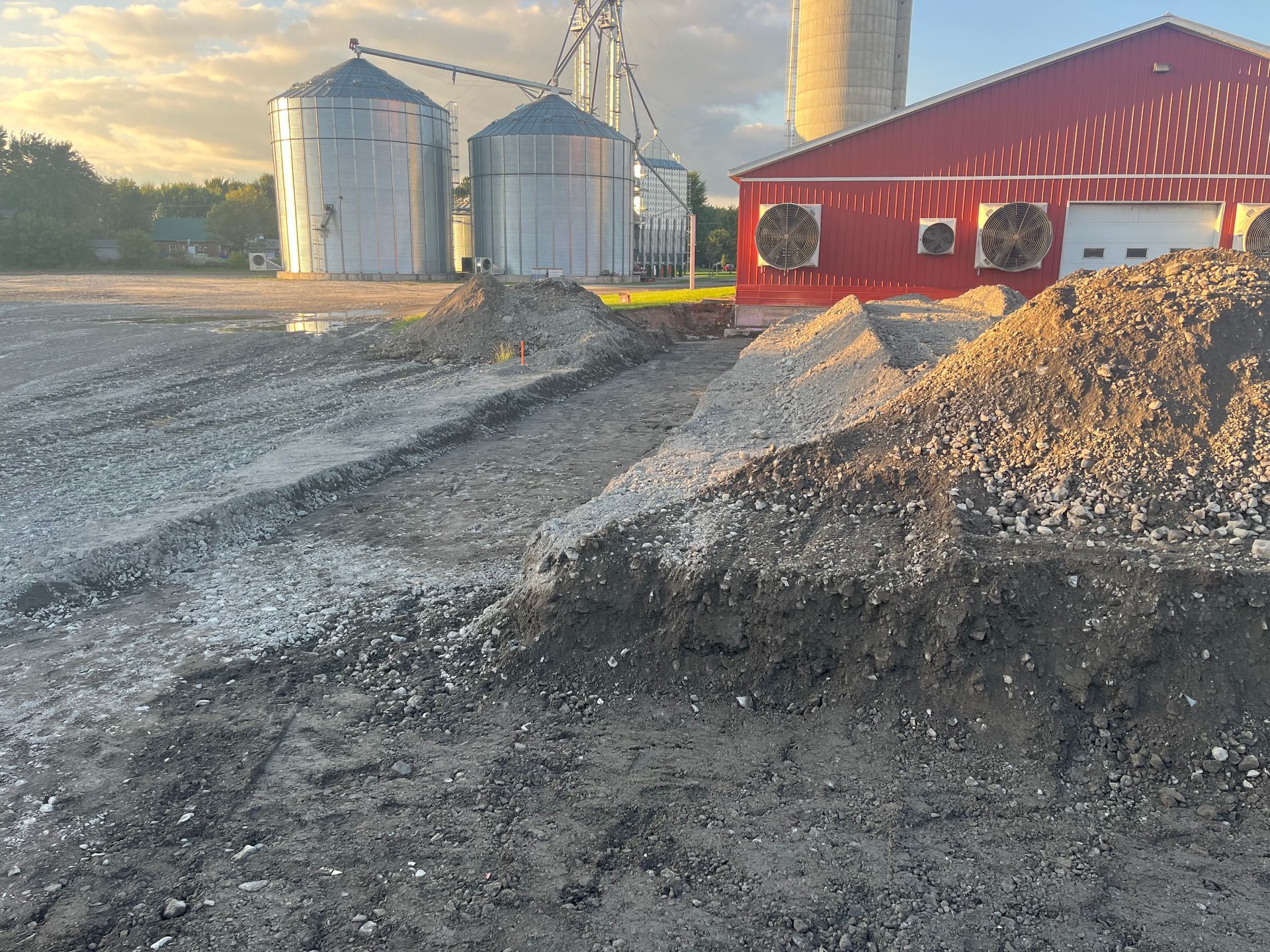 Allée de gravier en construction ; silos et bâtiment rouge en arrière-plan au crépuscule.