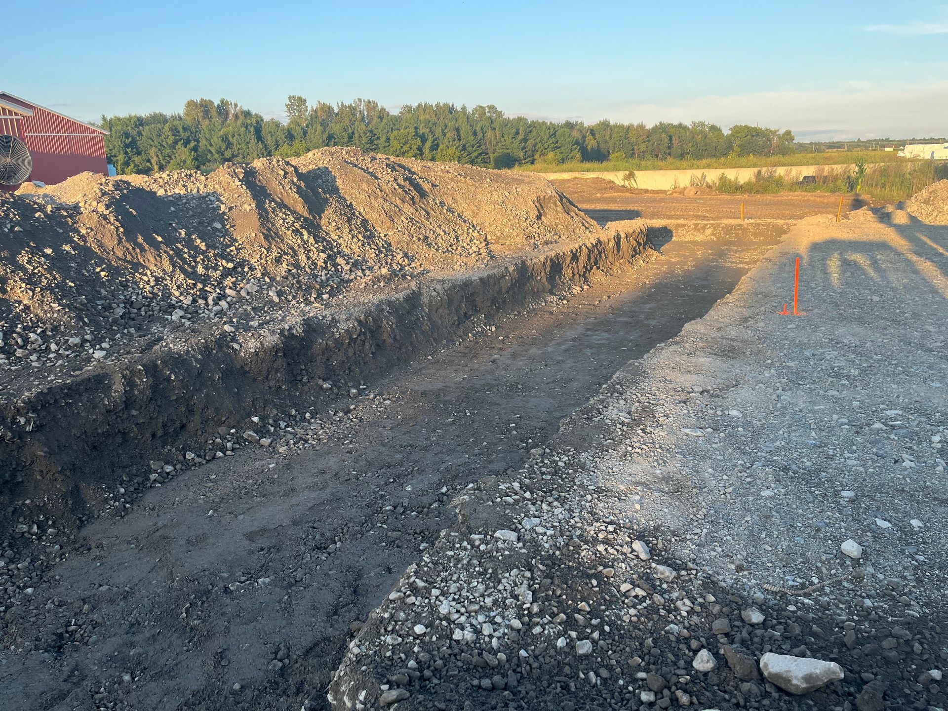 Chantier de construction avec des tranchées remplies de gravier et de terre. Tas de terre et arbres en arrière-plan.