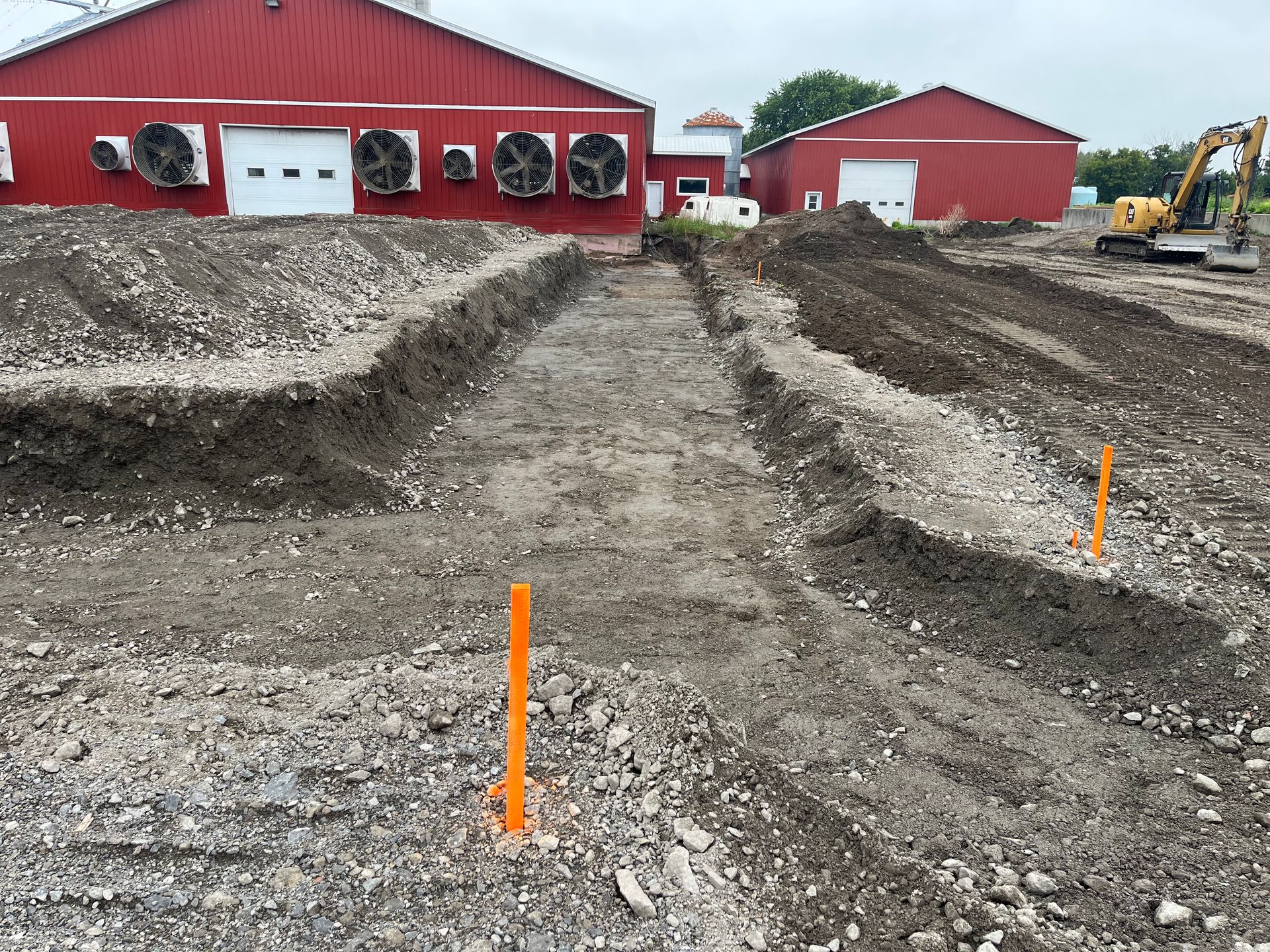 Des tranchées de terre ont été creusées devant des bâtiments agricoles rouges ; des balises orange et des engins de chantier sont visibles.