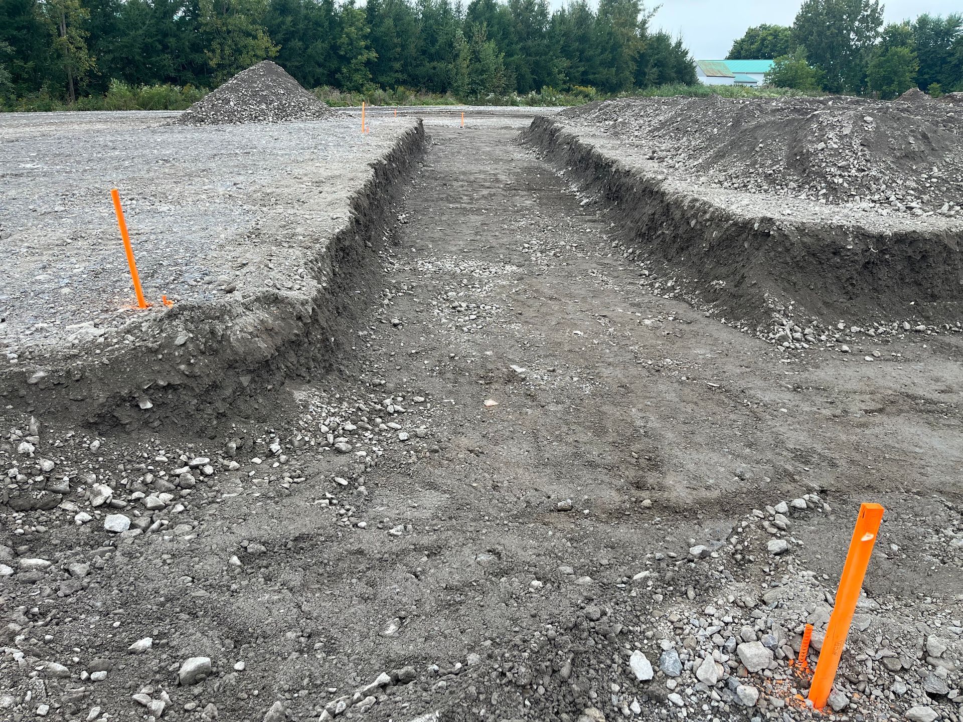 Un chantier de construction avec des tranchées creusées, du gravier, des tas de terre et des balises orange.