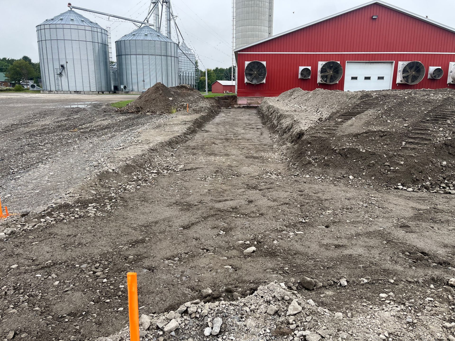 Chantier, tranchée creusée devant une grange rouge et des silos, tas de terre.