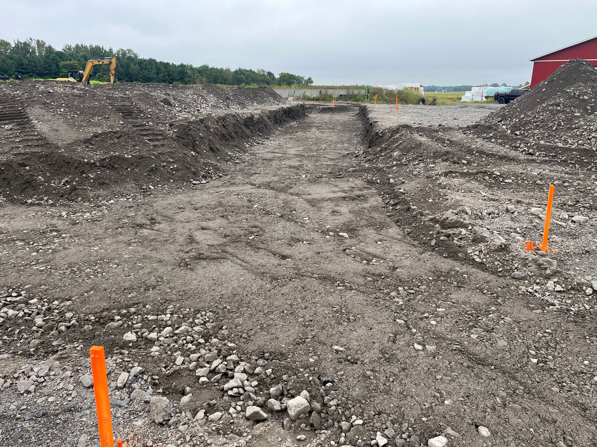 Chantier avec tranchées creusées dans un sol caillouteux. Tas de terre sur les côtés, balises orange présentes.