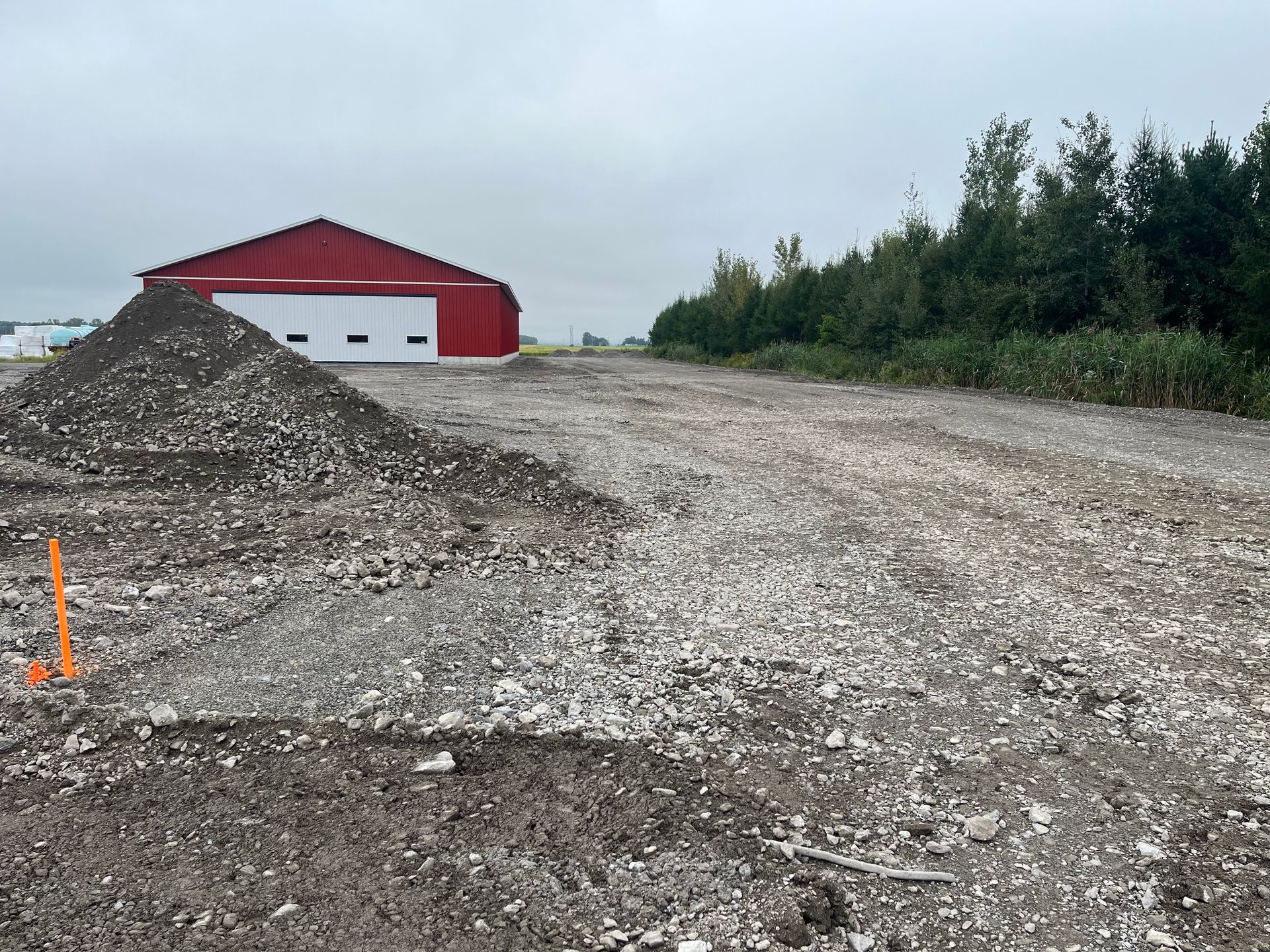 Terrain gravillonné avec un bâtiment rouge et des portes blanches, un monticule de gravier, des arbres à droite, ciel nuageux.
