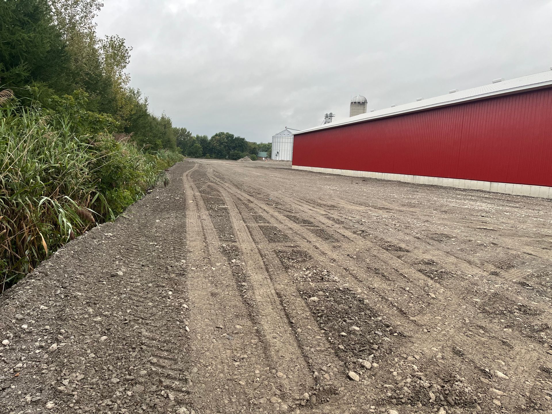 Chemin de gravier longeant une grange rouge par temps nuageux. La route est marquée par des traces de pneus et de la végétation pousse sur la gauche.
