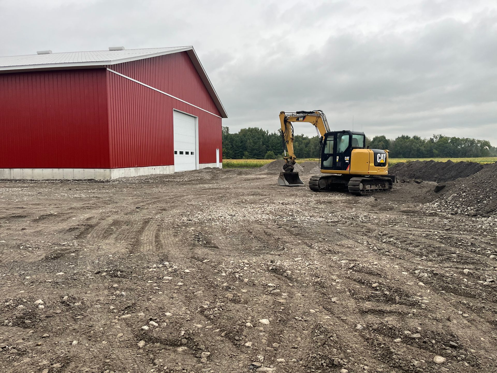 Une pelleteuse jaune est garée près d'une grange rouge. Le sol est recouvert de gravier. Ciel couvert.
