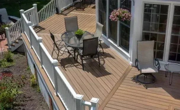 An elevated wooden deck with a glass-topped dining set and a single chair, framed by white railings next to a house.