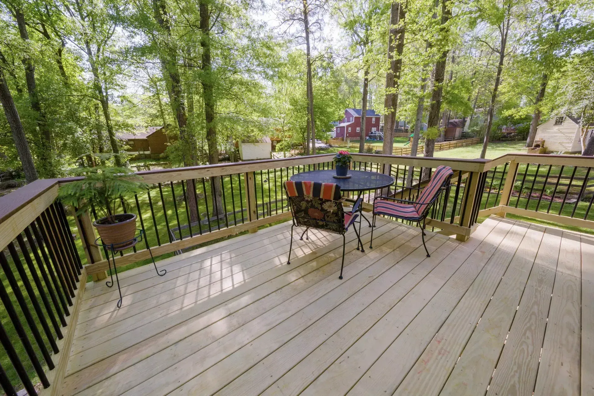 A wooden deck with a small round table and two chairs, overlooking a wooded yard with distant houses.