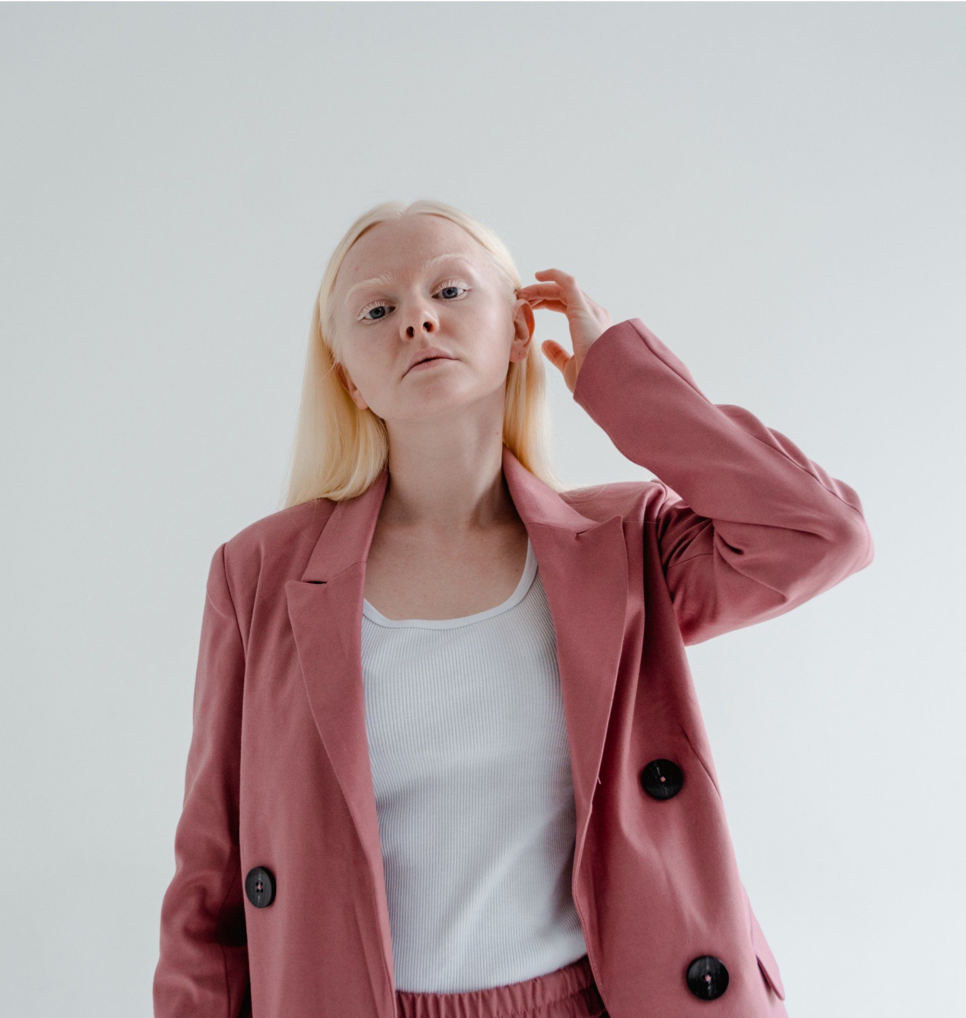 Woman in pink blazer and white top, hand near head, looking at the camera against a white backdrop.