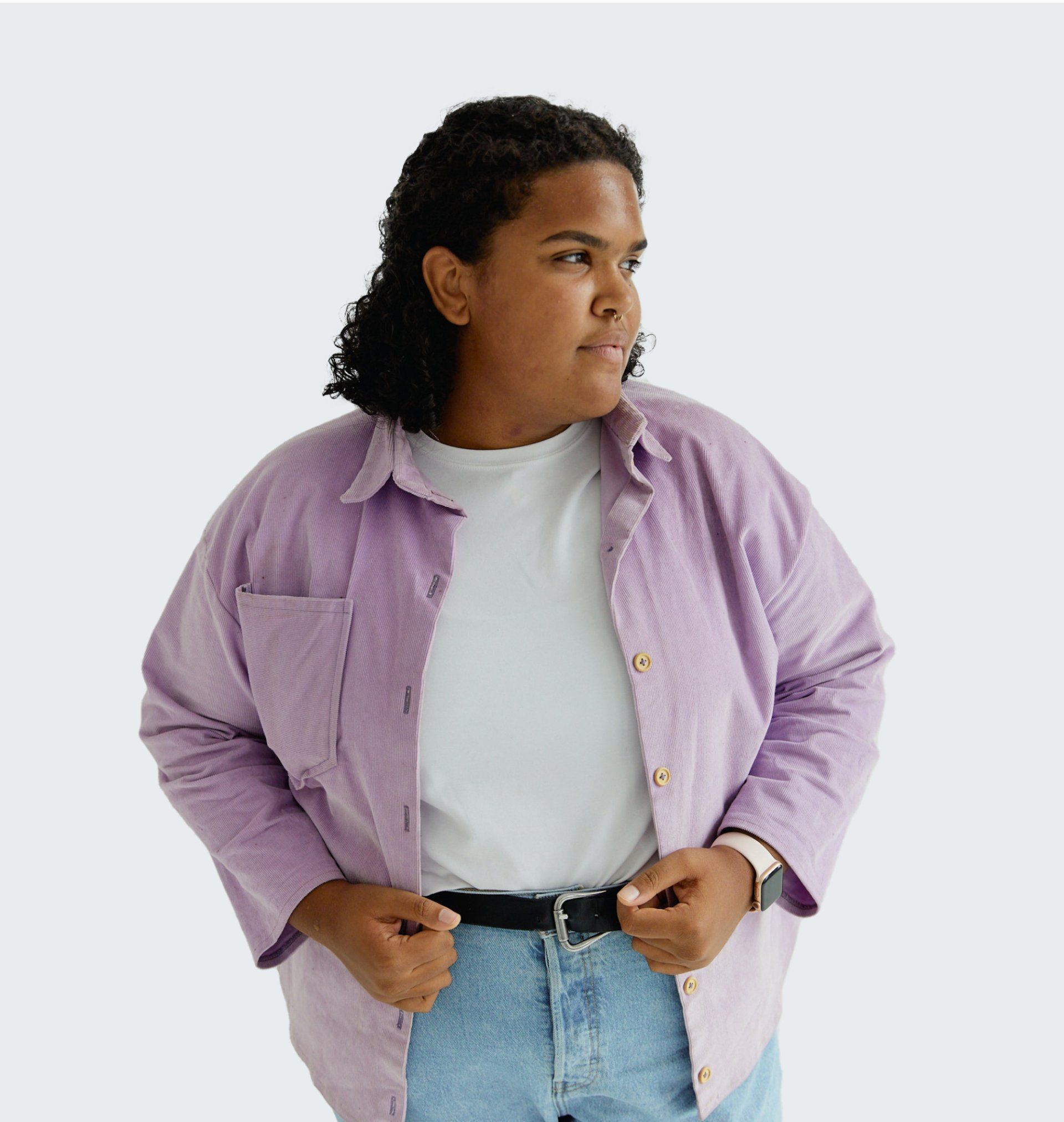 Person in a lilac shirt, white tee, and jeans looking to the side. Hands near belt, neutral expression.