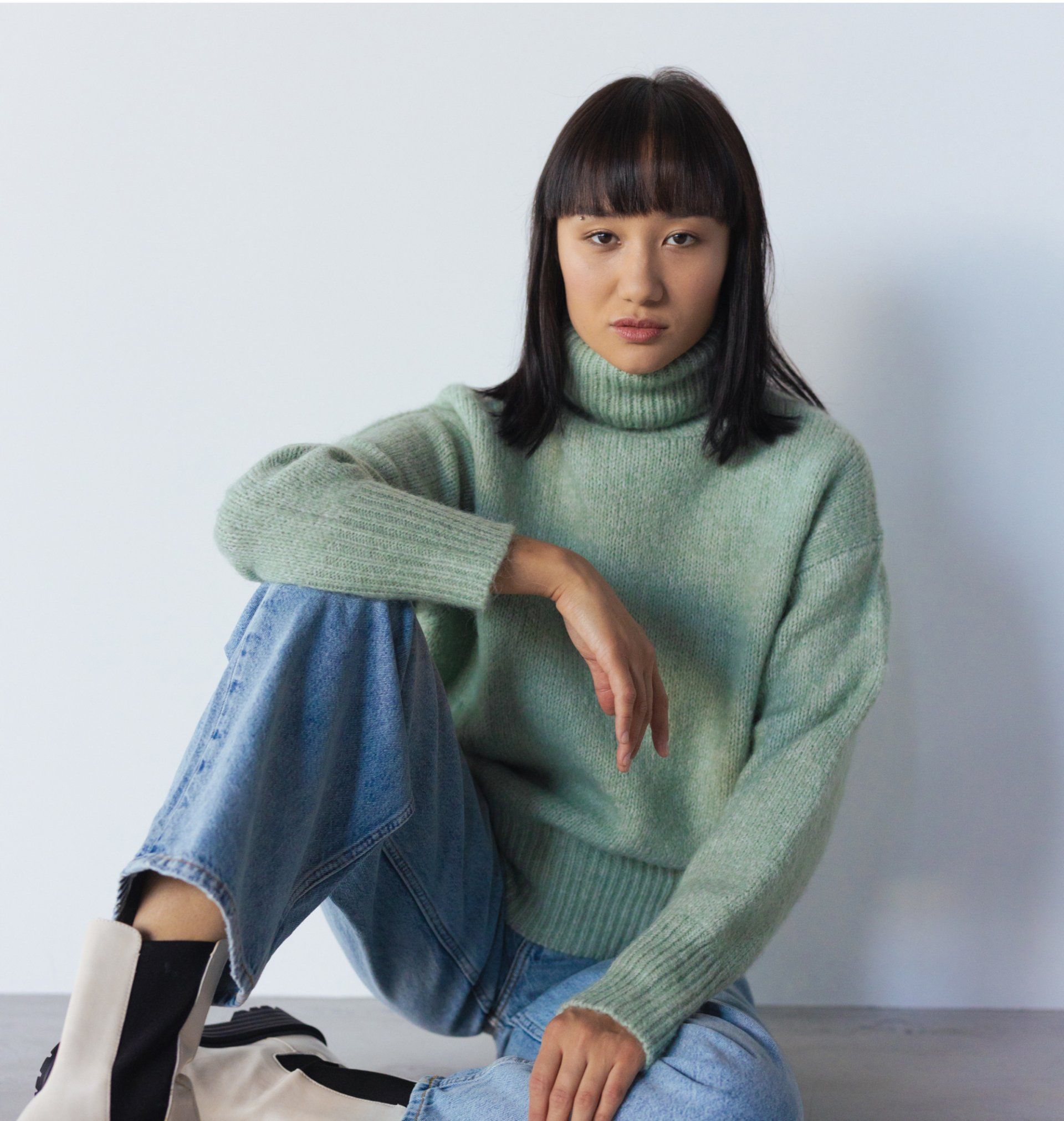 Person in green turtleneck pulls it up, partially covering face; seated, wearing jeans.