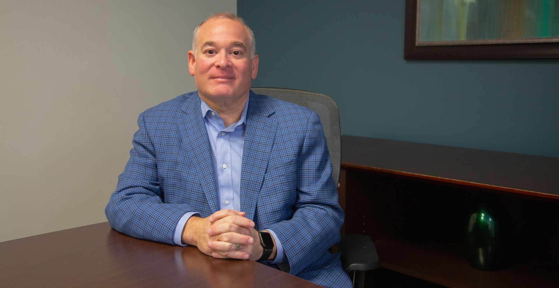 A man in a suit is sitting at a desk with his hands folded.
