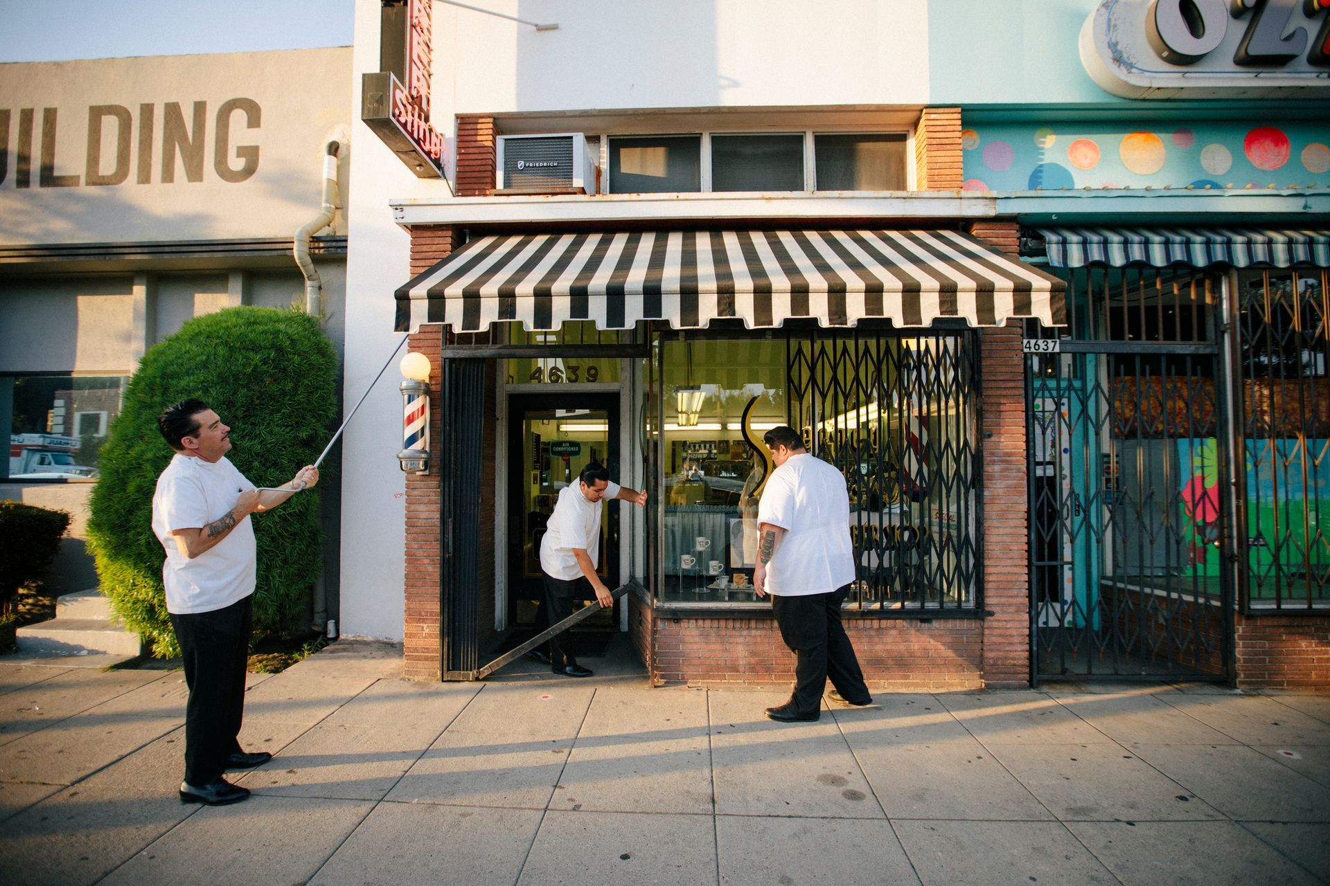 Three men at a barber shop, one holding pole sign, others enter, black & white awning, sunny exterior.