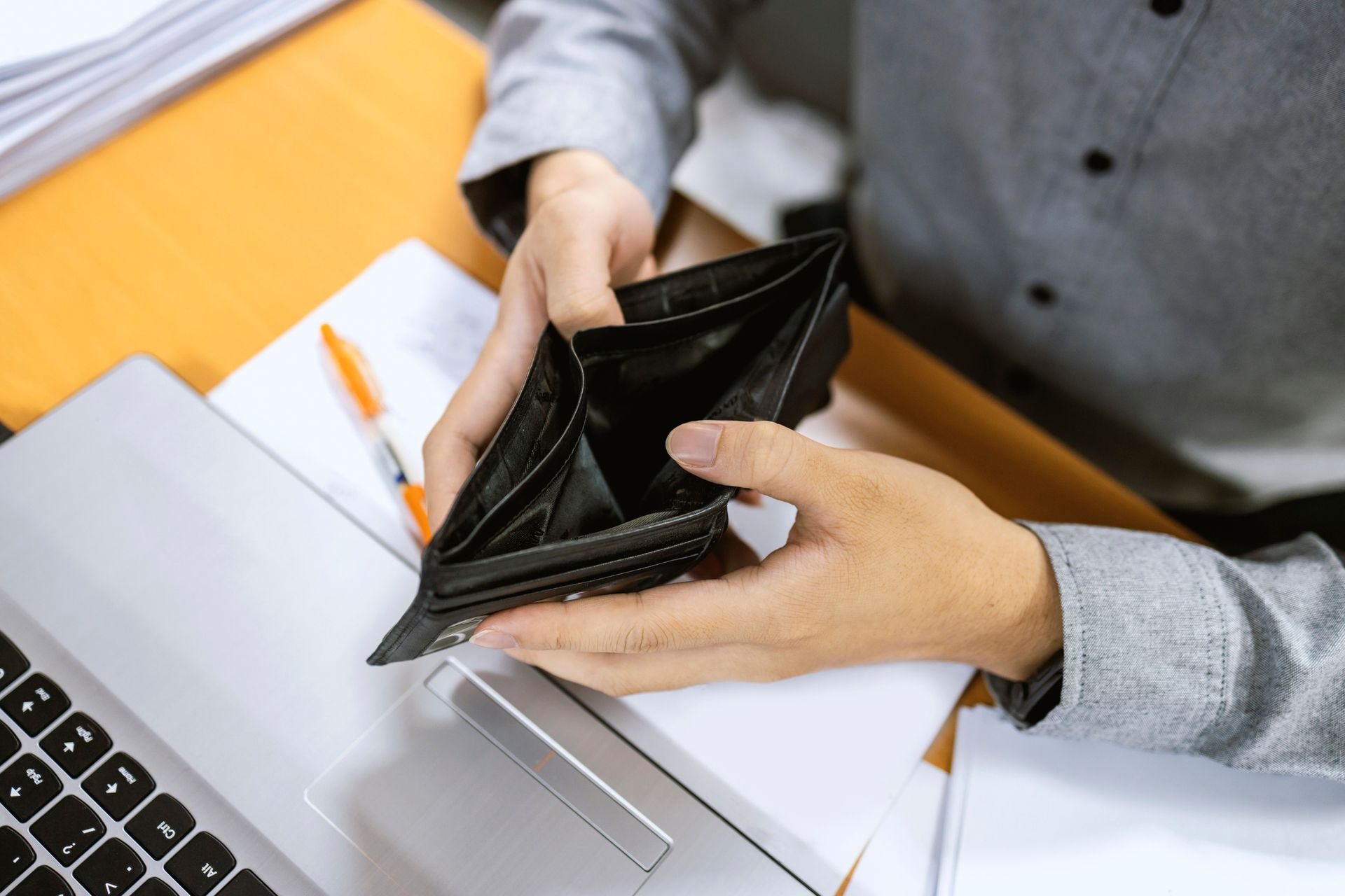 Person holding an empty black wallet, near a laptop and paperwork, conveying financial hardship.