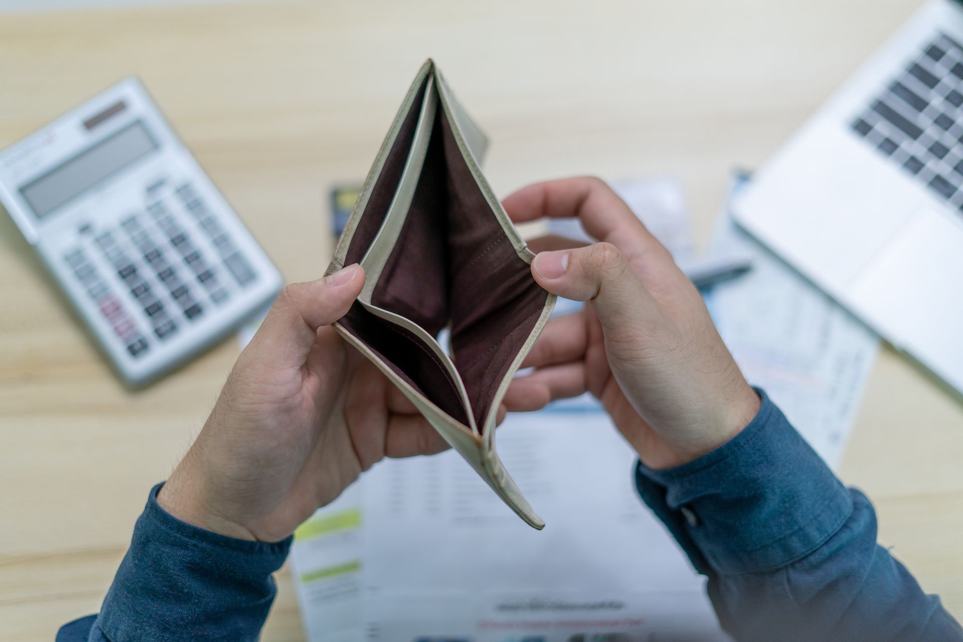 Hands holding an empty wallet, with a calculator, laptop, and bills in the background, indicating financial difficulties.