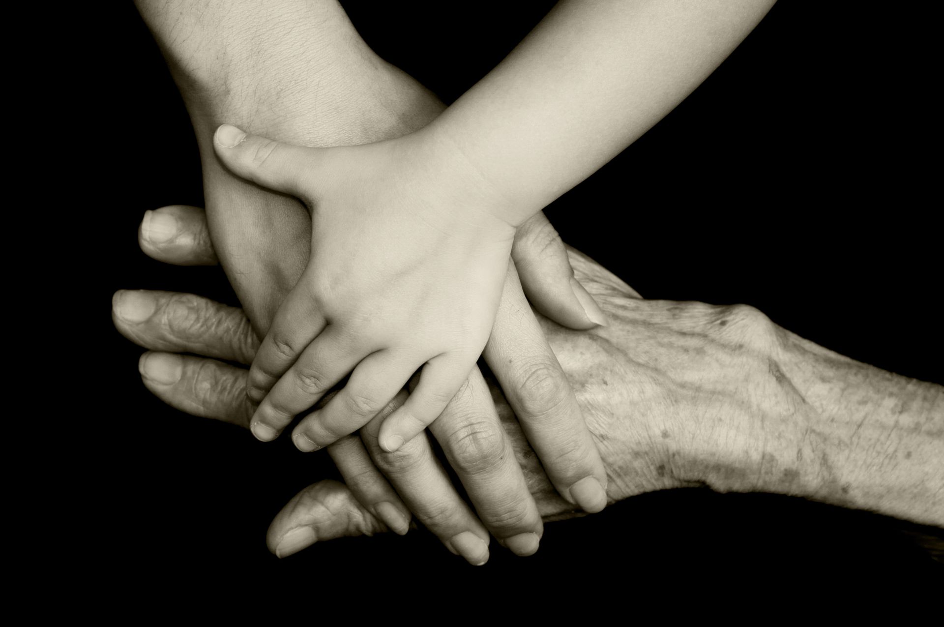Hands of different ages, child's hand on top of an adult's, both resting on an older person's hand.