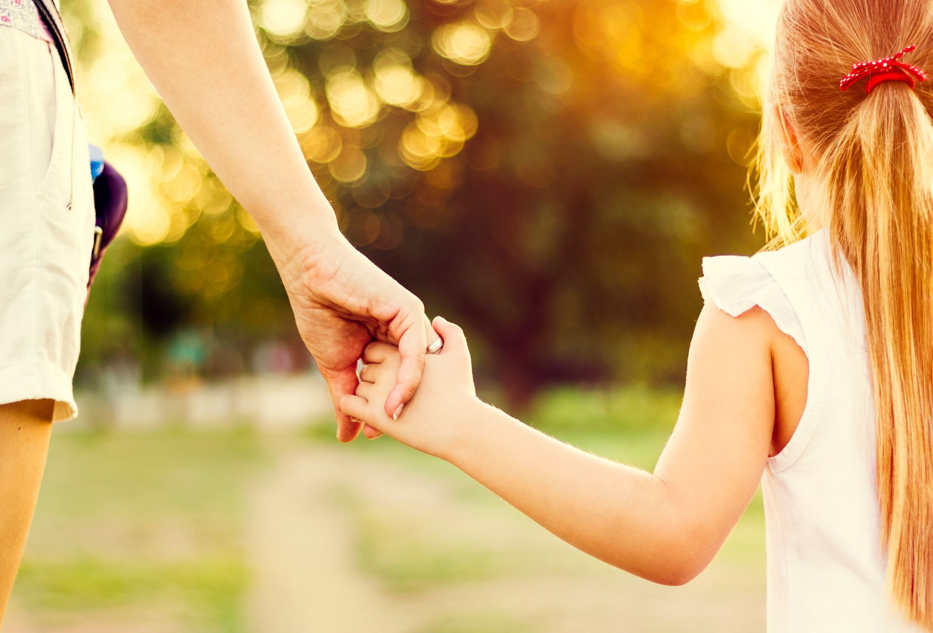 Mother and child holding hands, walking outdoors in sunlight.