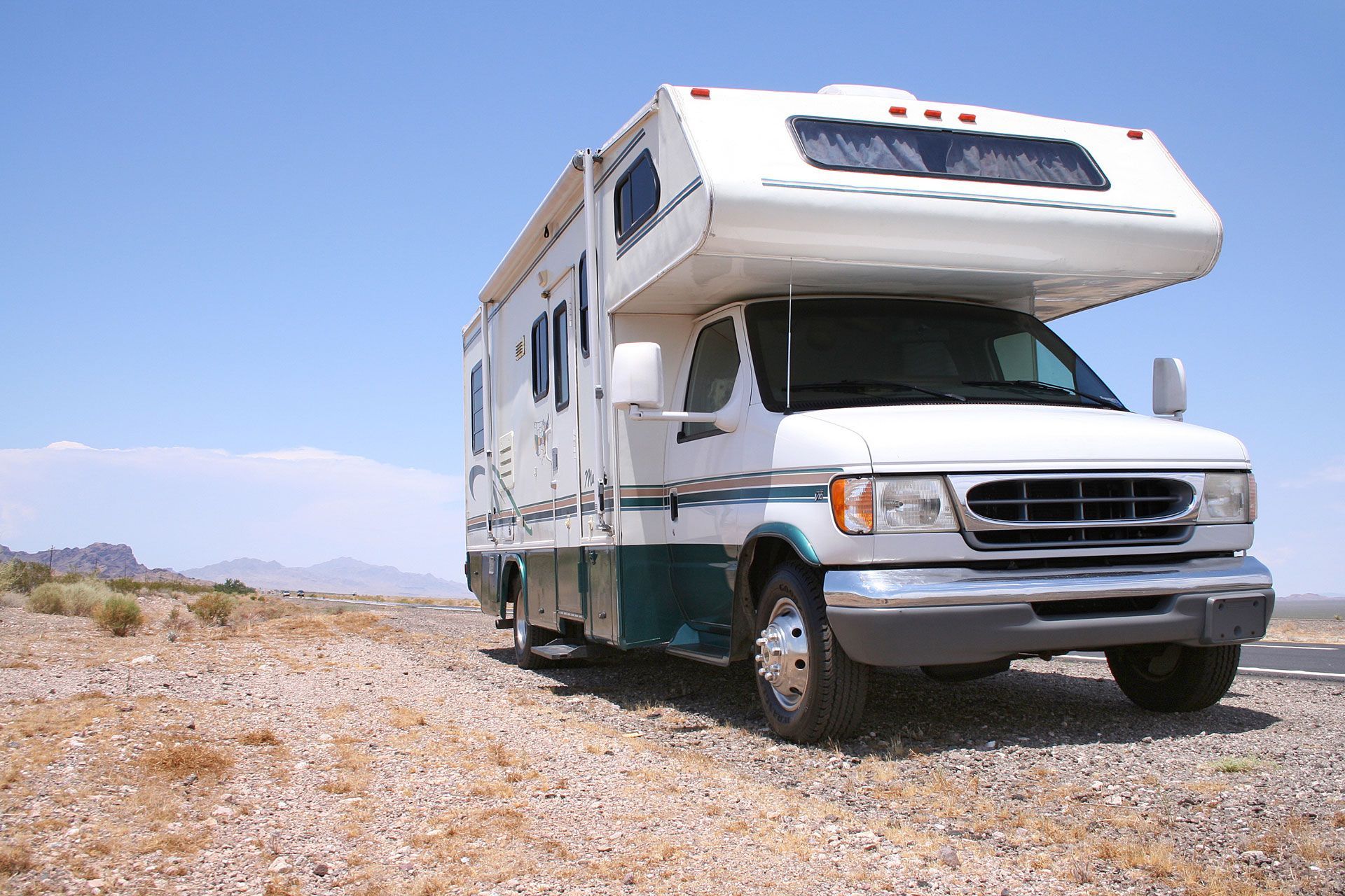 A white and teal motorhome parked on a gravel roadside under a clear blue sky in a desert landscape.