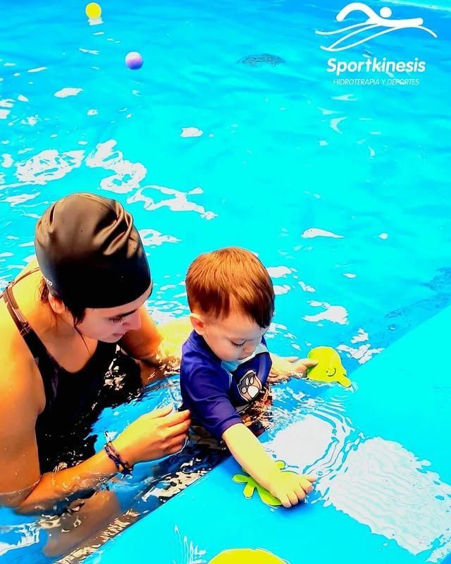 Una mujer con gorro de baño ayuda a un niño pequeño en la piscina; ambos intentan alcanzar sus juguetes.