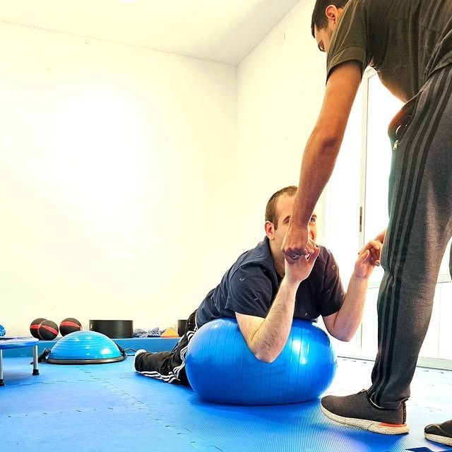 Hombre sobre una pelota de ejercicio azul con un entrenador ayudándolo en un gimnasio.