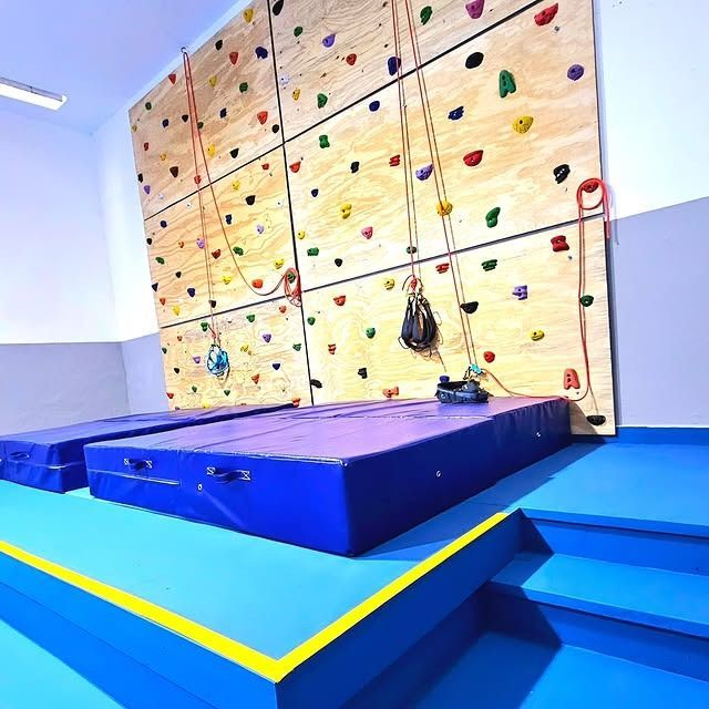 Muro de escalada interior con presas de colores, cuerdas y colchonetas de seguridad; detalles en azul y amarillo.