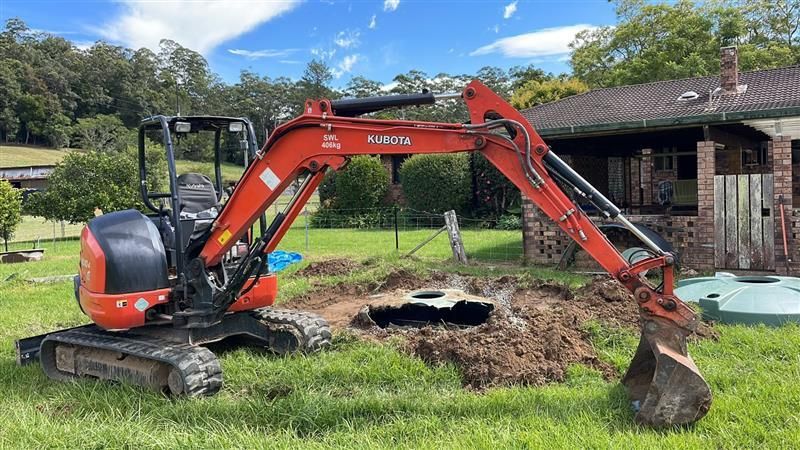 A Red Excavator Parked on Grass — Tim Larkin Plumbing and Excavations in Lake Cathie, NSW