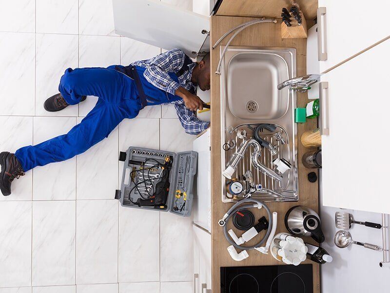 A Plumber Is Laying On The Floor Fixing A Sink In A Kitchen — Tim Larkin Plumbing and Excavations in Lake Cathie, NSW