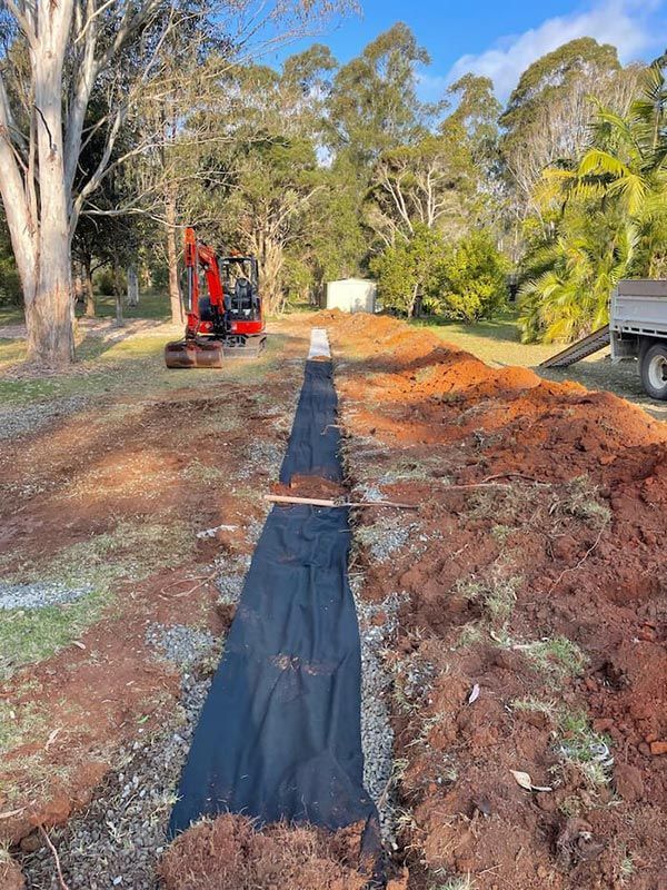 A Red Excavator Is Digging A Land In The Dirt Next To A Truck For Pipeline — Tim Larkin Plumbing and Excavations in Wauchope, NSW