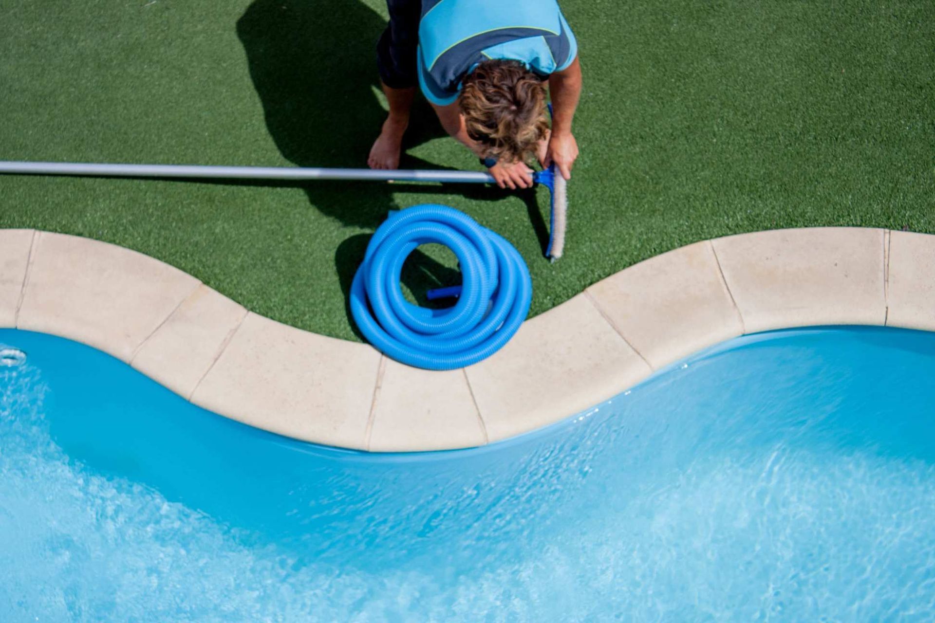 pool owner in the process of cleaning the artificial turf pool surround with a broom and a garden hose