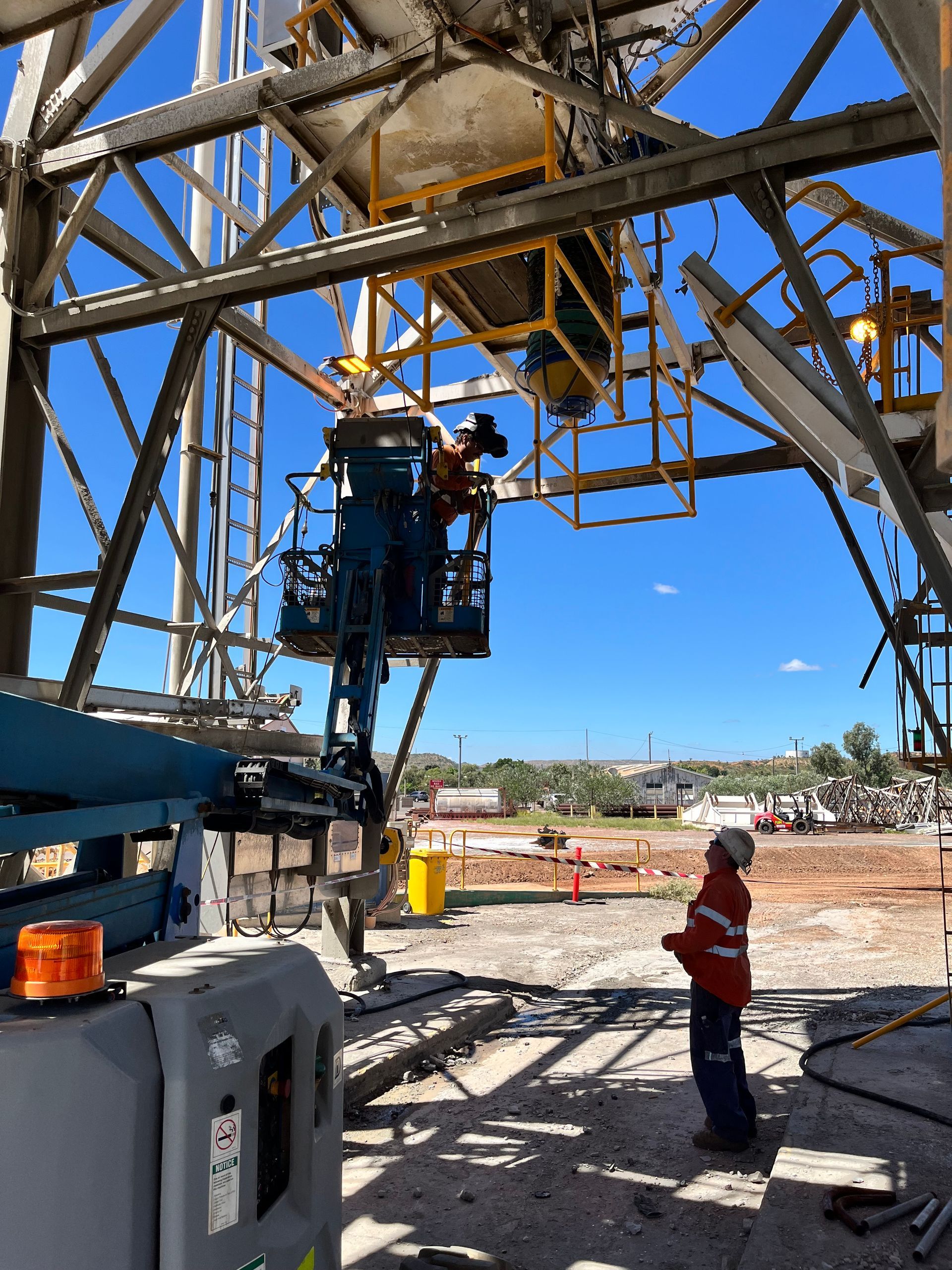 A worker in a high-visibility uniform watches a colleague in an aerial lift working on the metal structure of a rig.