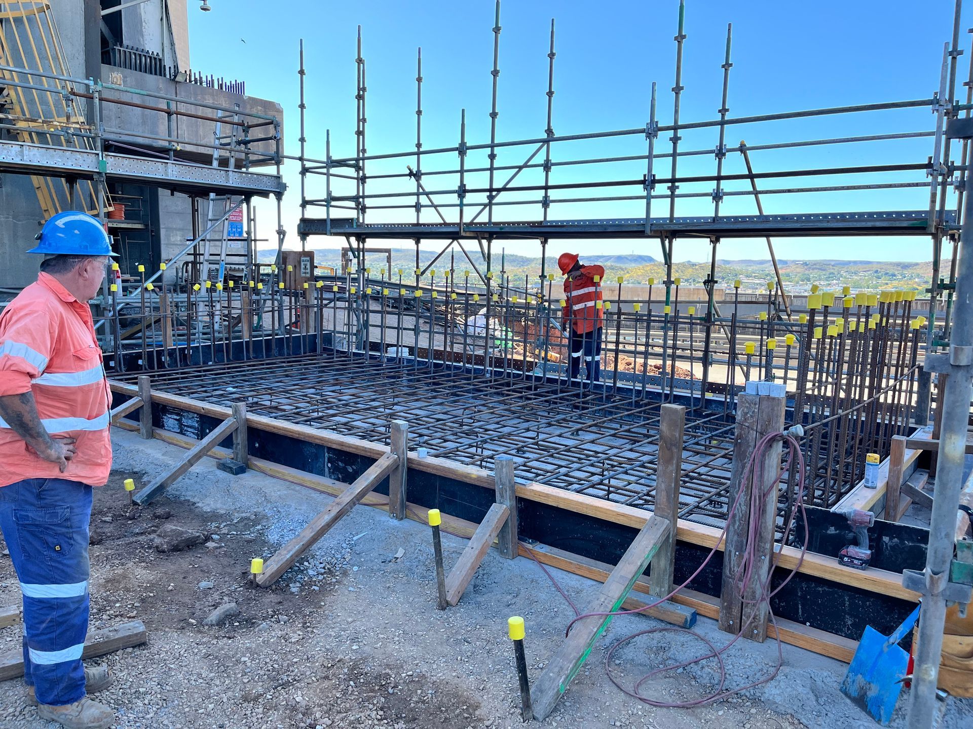 Two construction workers in high-visibility gear on a work site with scaffolding and steel reinforcement for a concrete slab.