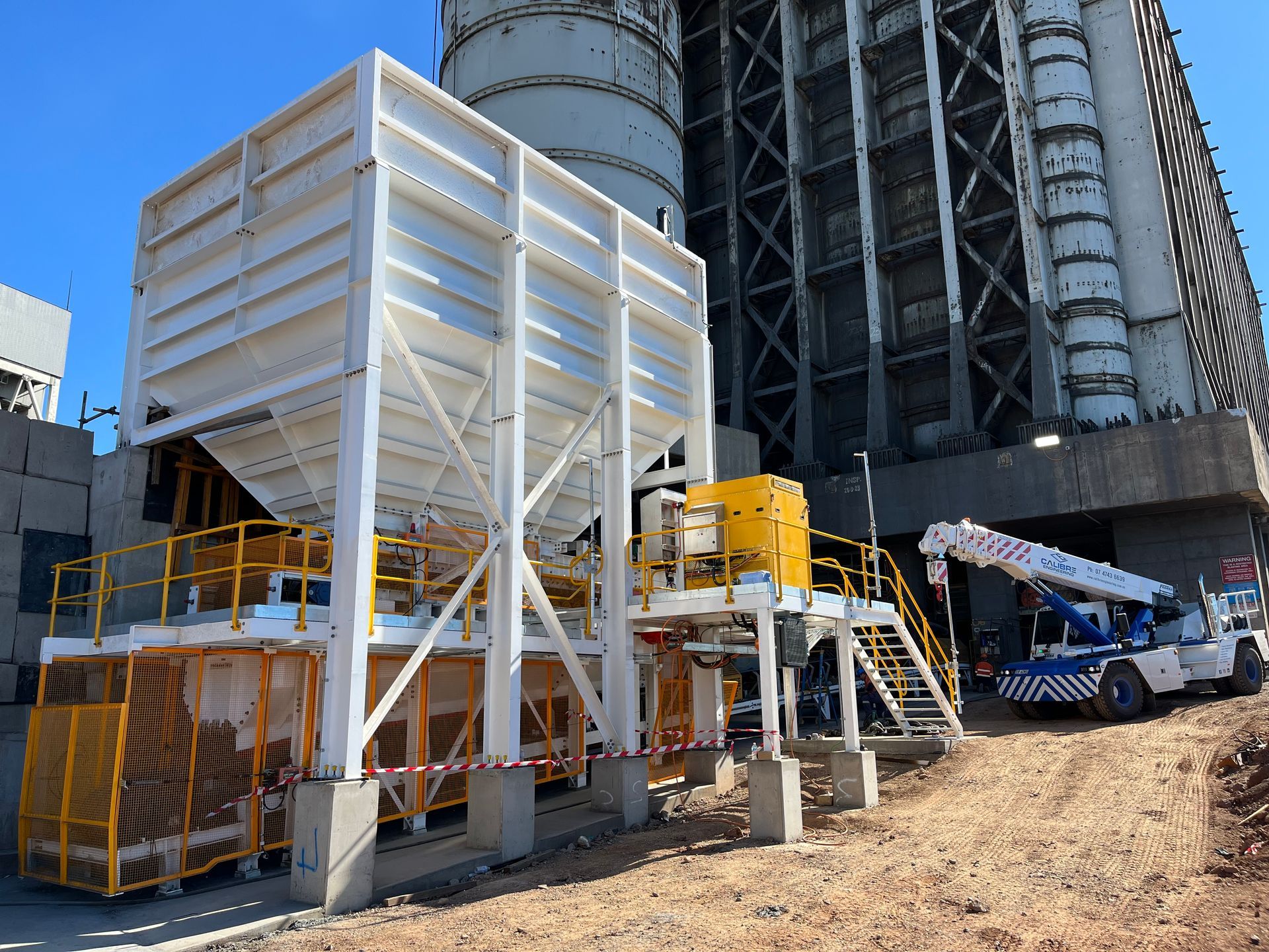 A large white industrial hopper stands on a concrete foundation next to a tall, weathered industrial building and crane.