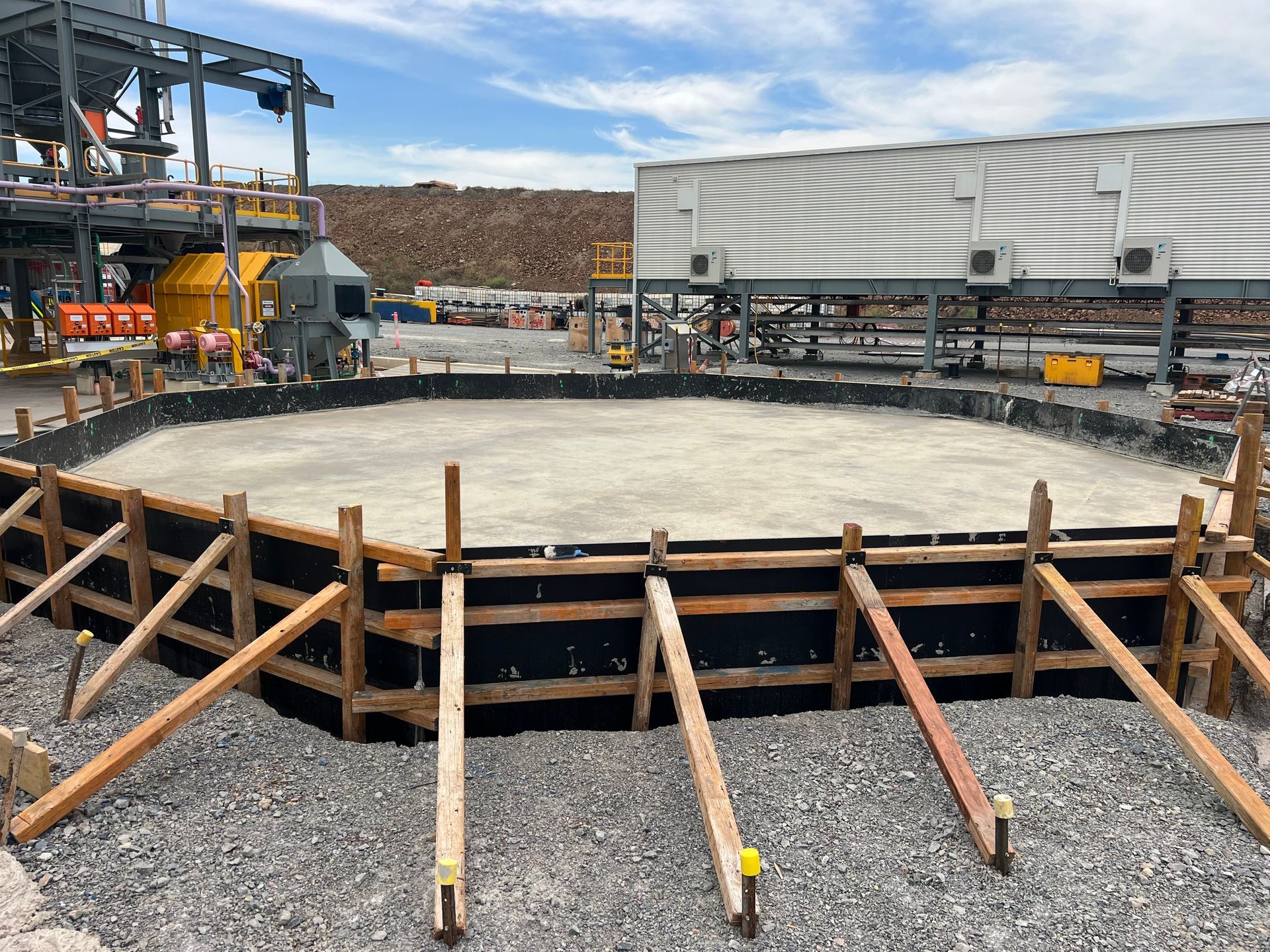 A circular concrete foundation under construction, surrounded by wooden formwork and angled support beams at a site.
