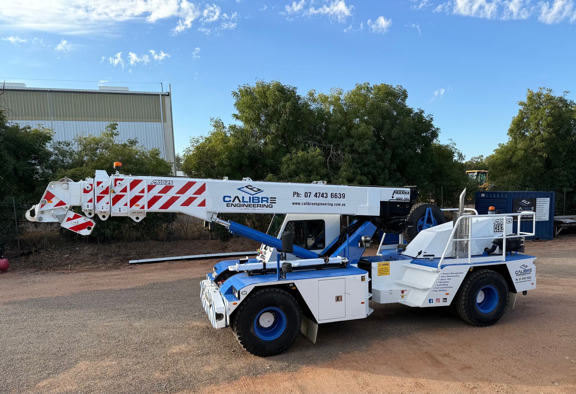 A white and blue industrial pick-and-carry crane parked on a gravel lot under a clear blue sky.