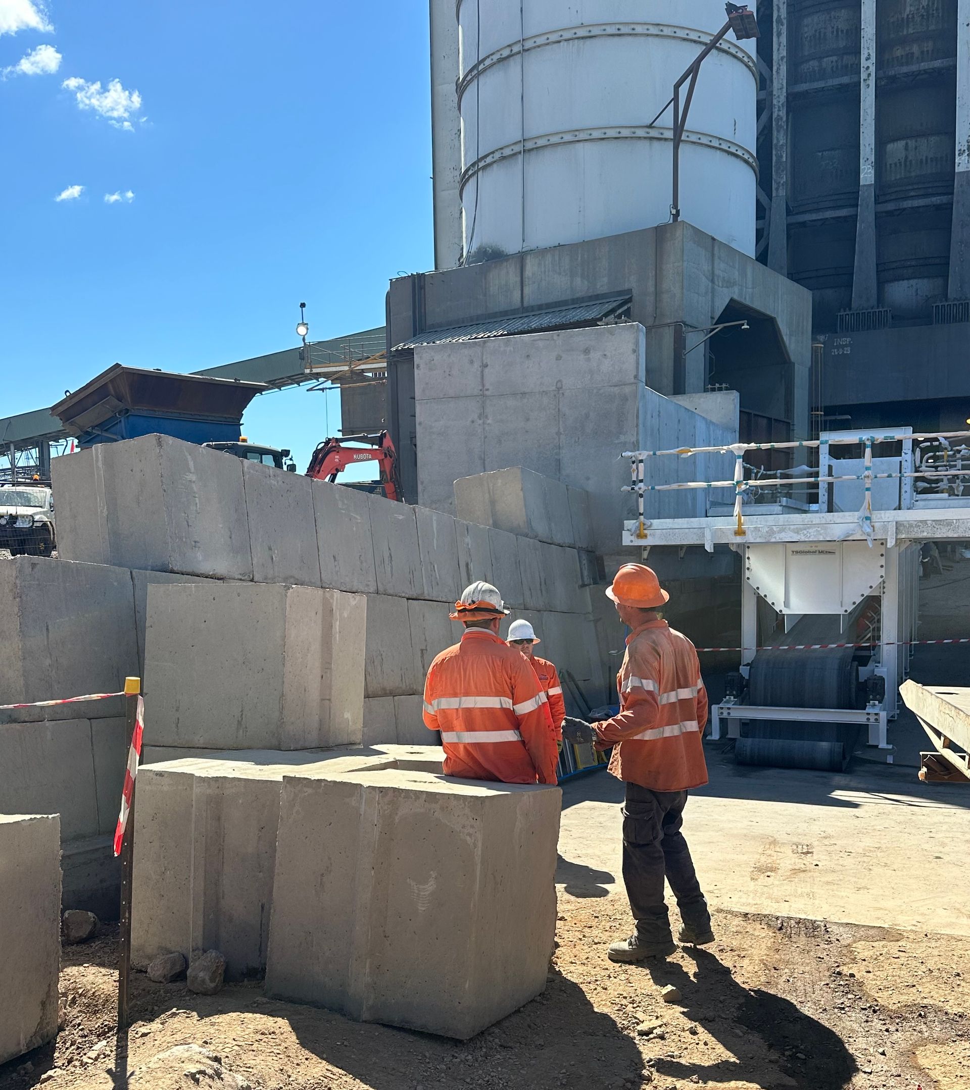 Three workers in bright orange safety gear stand together on a gravel site in front of large concrete blocks and a silo.