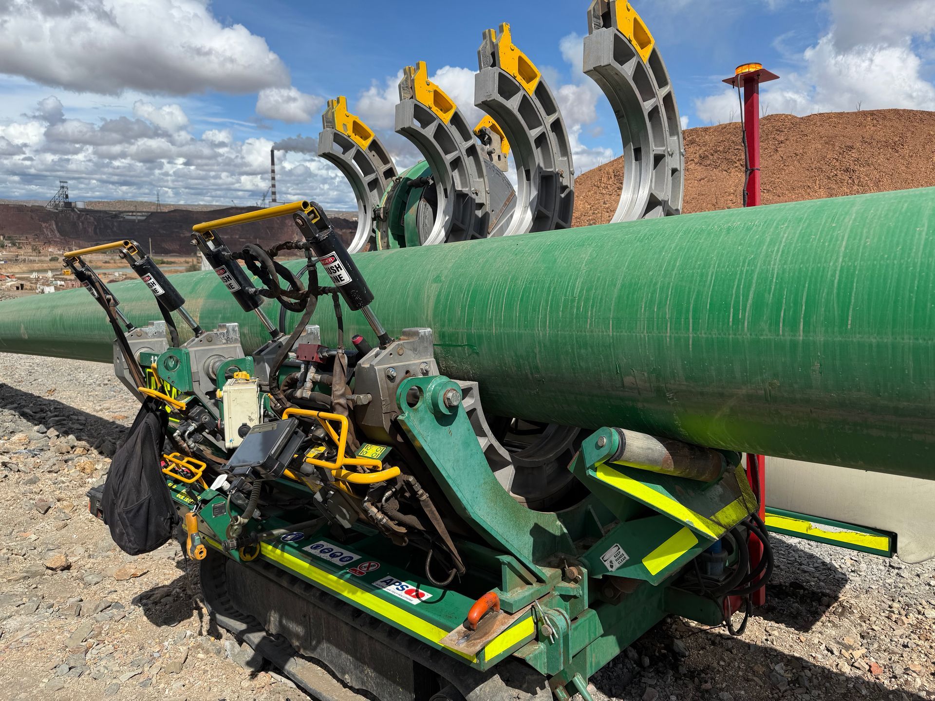 A green pipeline welding machine sits on rocky ground under a blue sky, holding a large green pipe section.