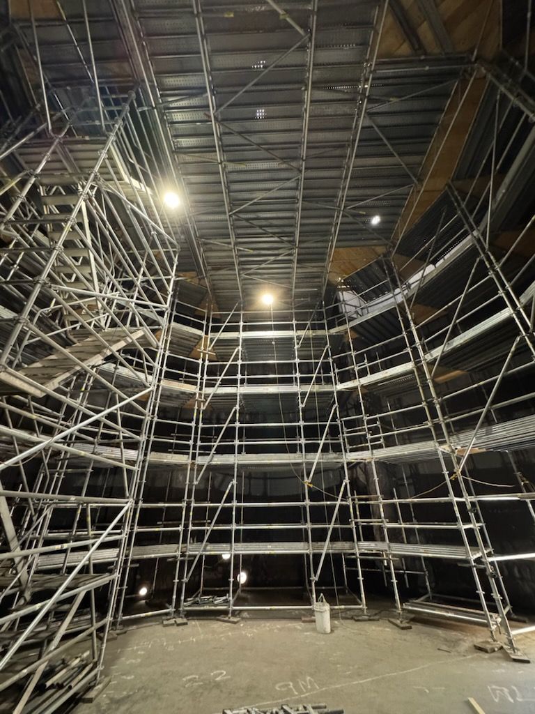 A multi-level metal scaffold structure set up in a large indoor space under high-mounted lights.