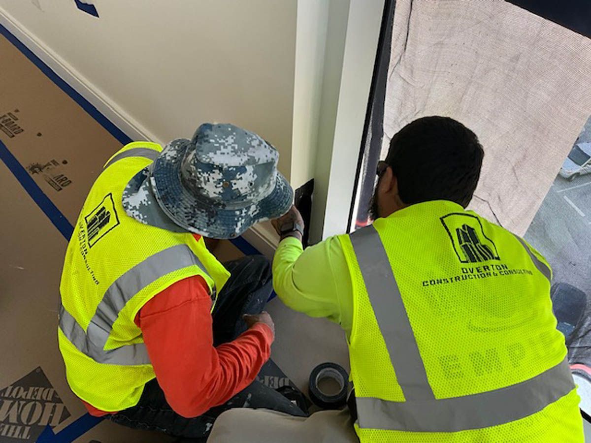 Two construction workers wearing yellow vests and hats are working on a window.