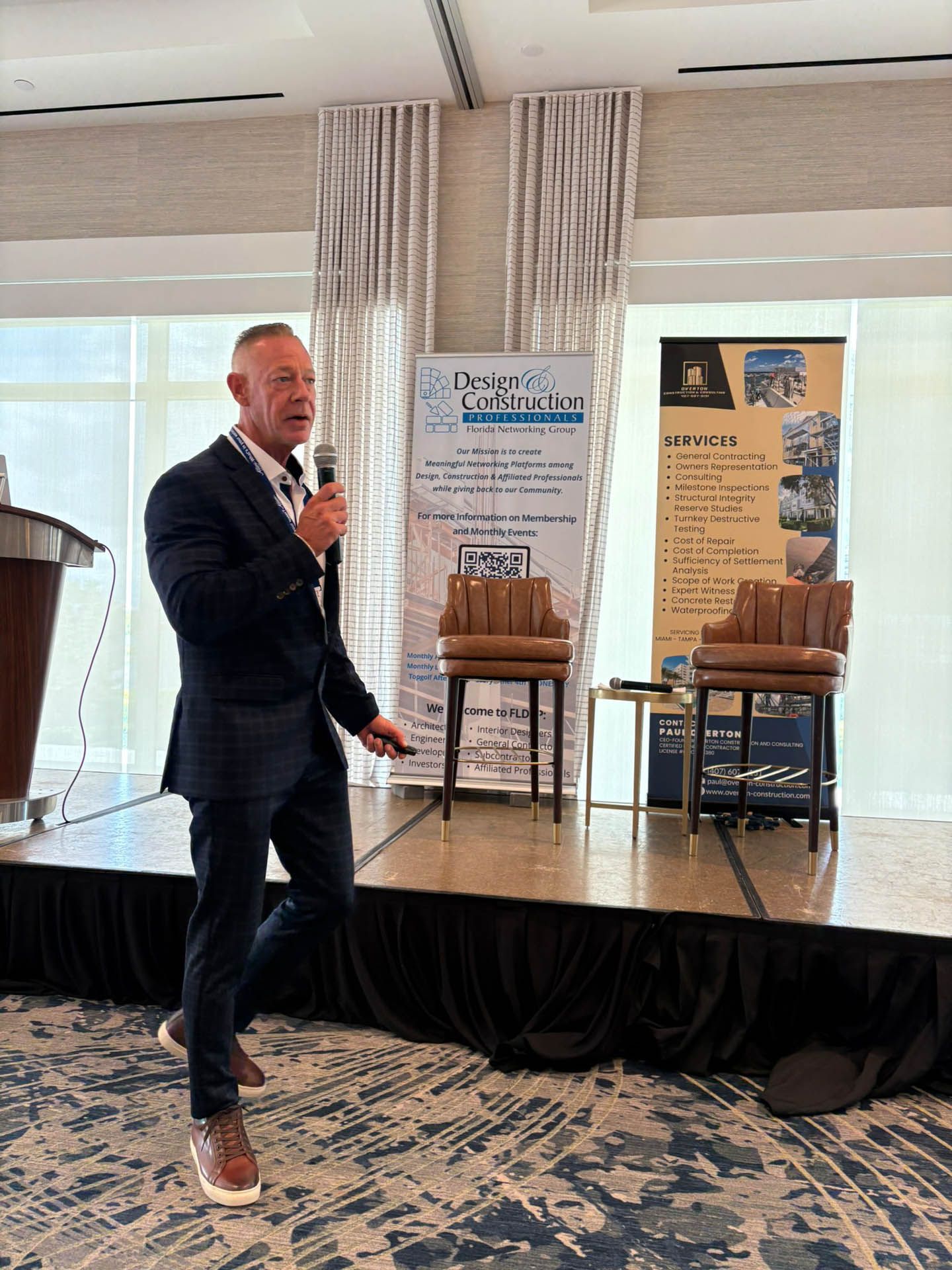 Man in suit speaks at a conference, holding a microphone, on a stage with two chairs.