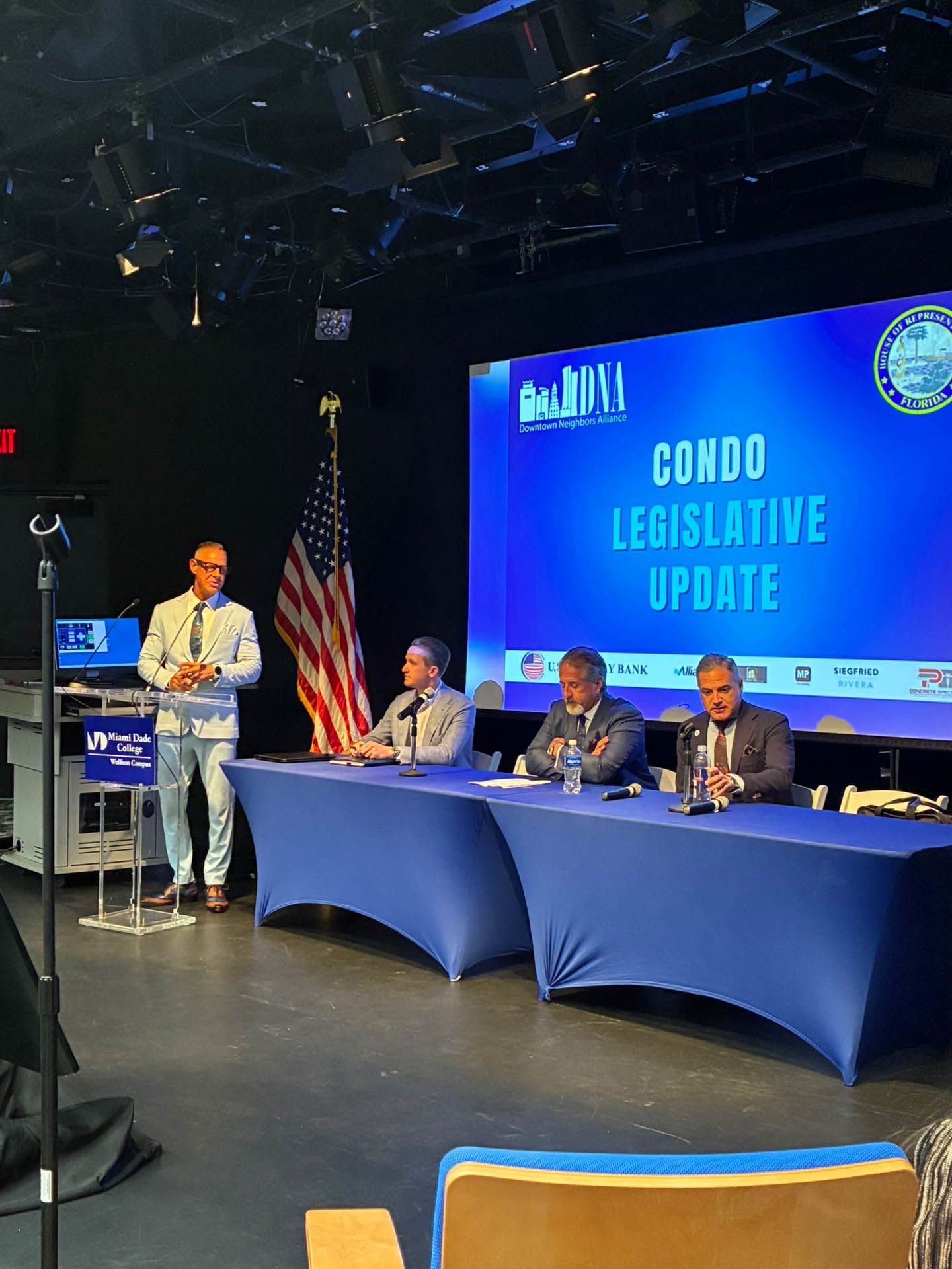 Four men at a table, discussing legislative update. Blue backdrop reads “CONDO LEGISLATIVE UPDATE”. American flag present.