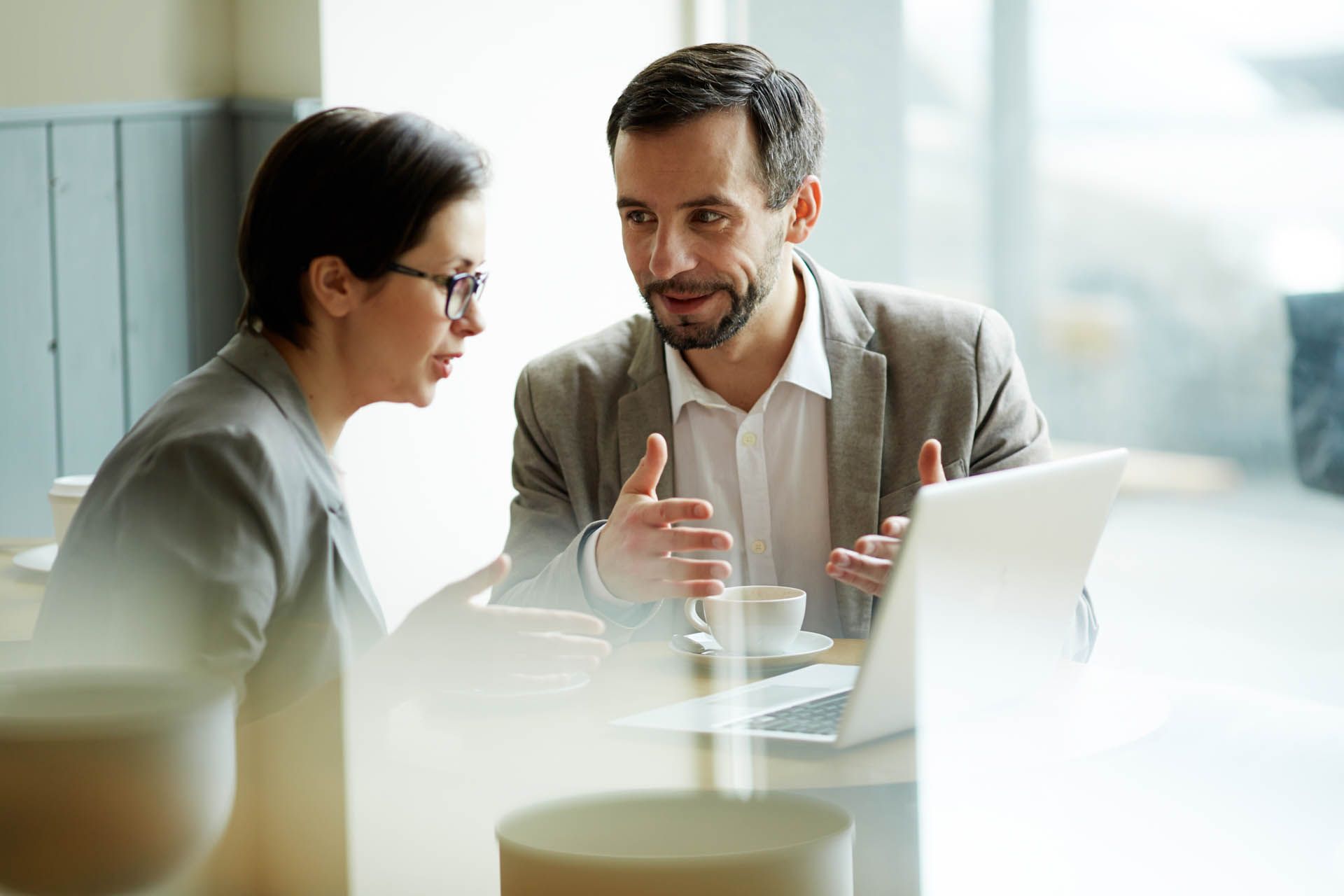 Two businesspeople at a table with a laptop, discussing and gesturing.