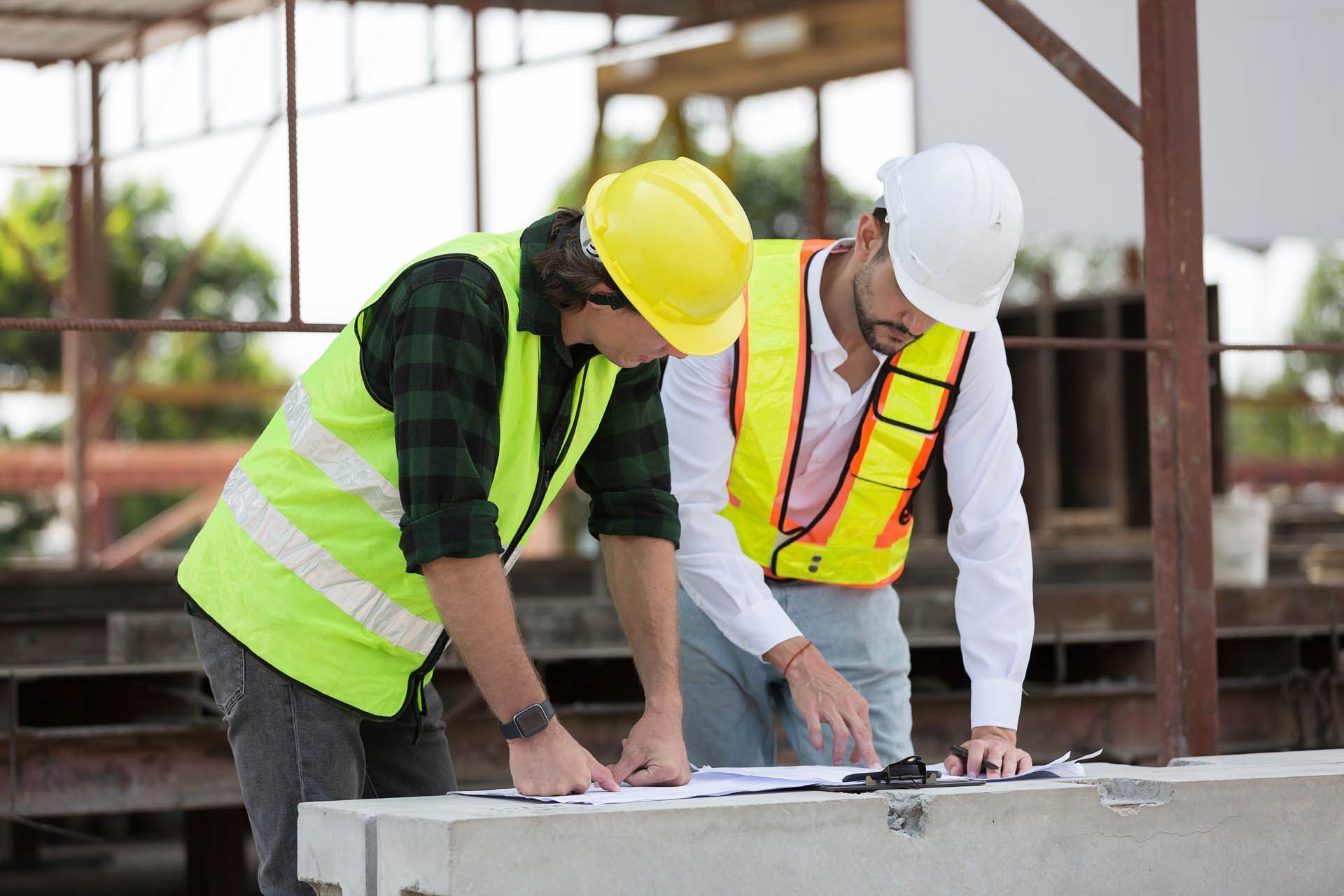 Two construction workers in vests and helmets examining blueprints outdoors.