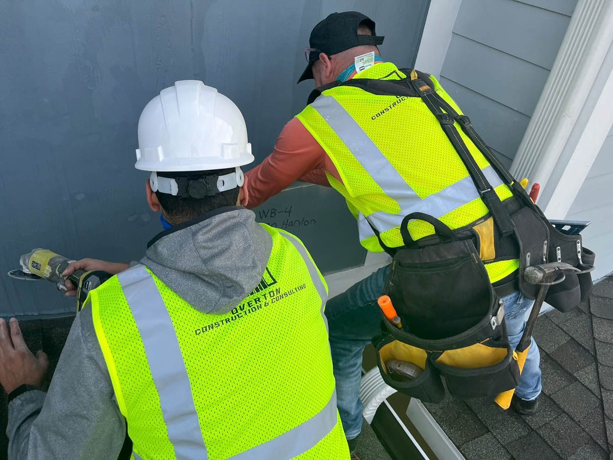 Two construction workers wearing yellow vests and hard hats are working on a house.