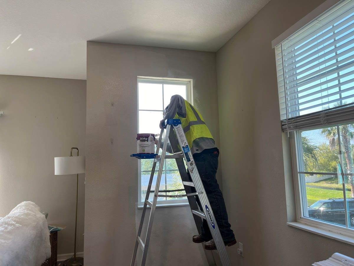 A man is standing on a ladder in front of a window in a bedroom.