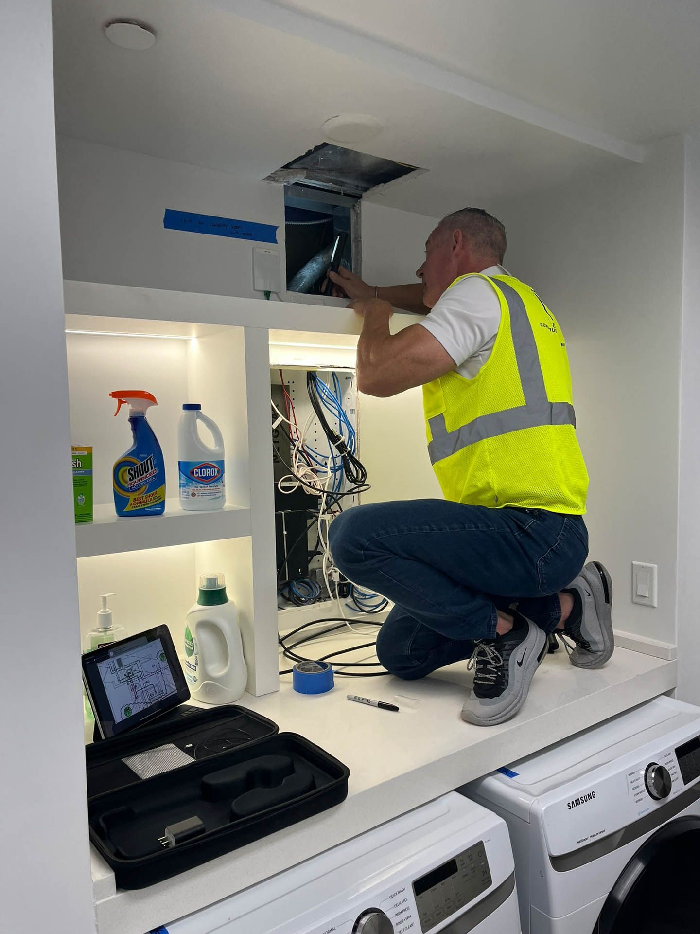 A man in a yellow vest is working on a washing machine in a laundry room.