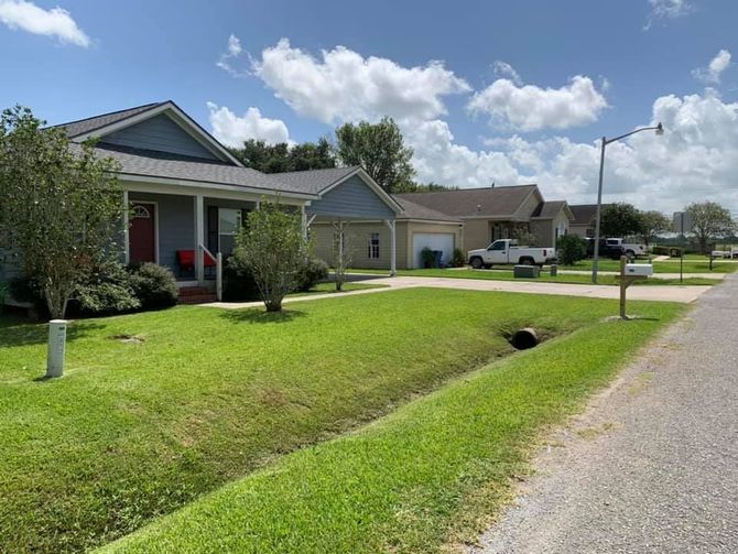 A row of suburban houses with gray siding and roofs, green lawns, and a ditch in the foreground under a sunny sky.
