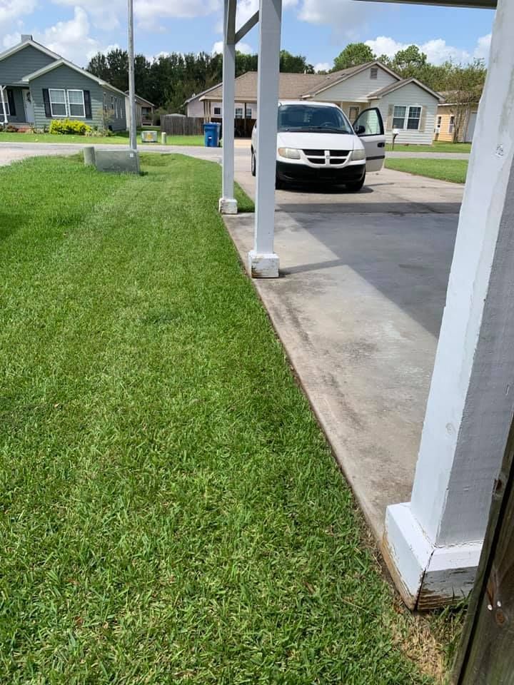 A grassy lawn bordered by a concrete driveway under a white-pillared carport, with a white car parked in the distance.