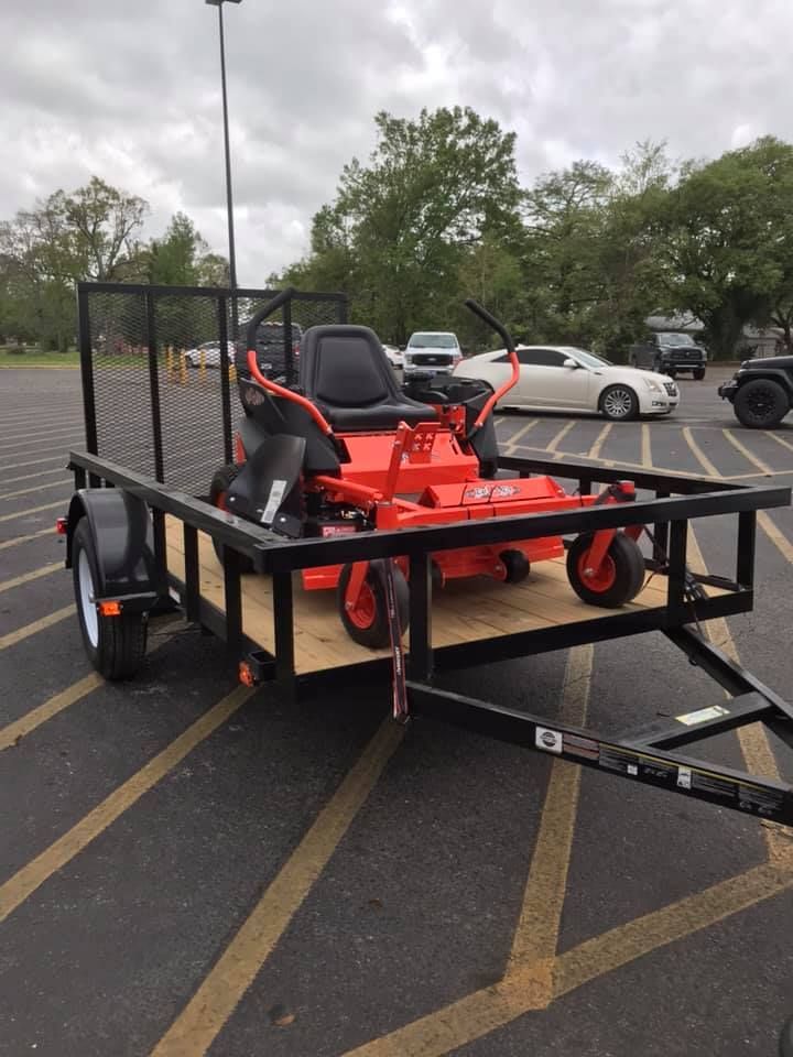 An orange zero-turn mower is secured on a black utility trailer in a parking lot on a cloudy day.