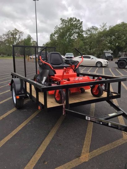 A bright orange zero-turn mower is strapped onto a black utility trailer parked in a large parking lot.