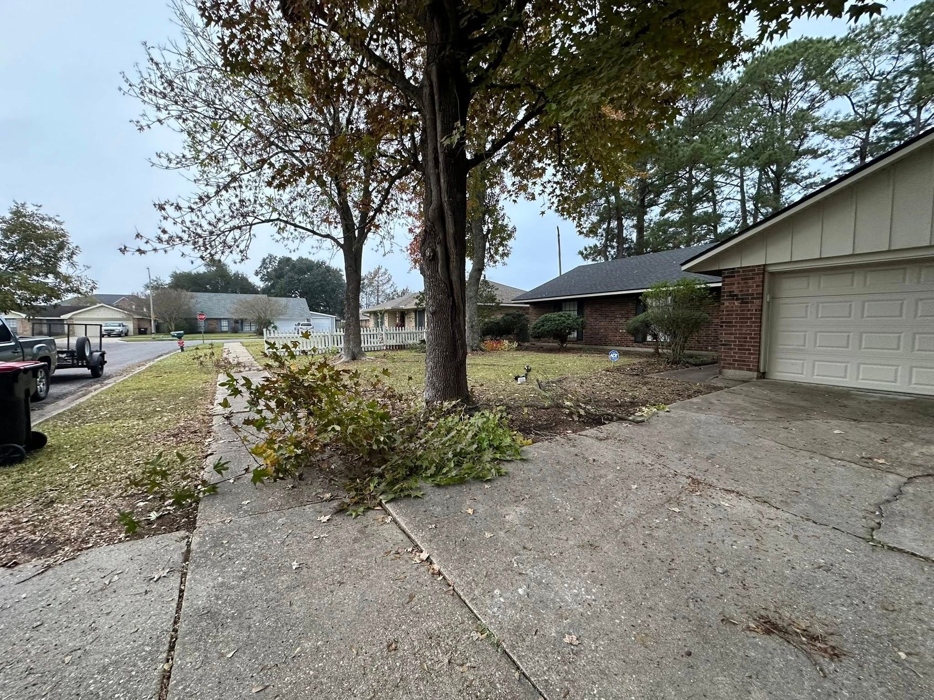 A concrete driveway leads to a brick house on a cloudy day, with fallen branches in the grass and a parked truck nearby.