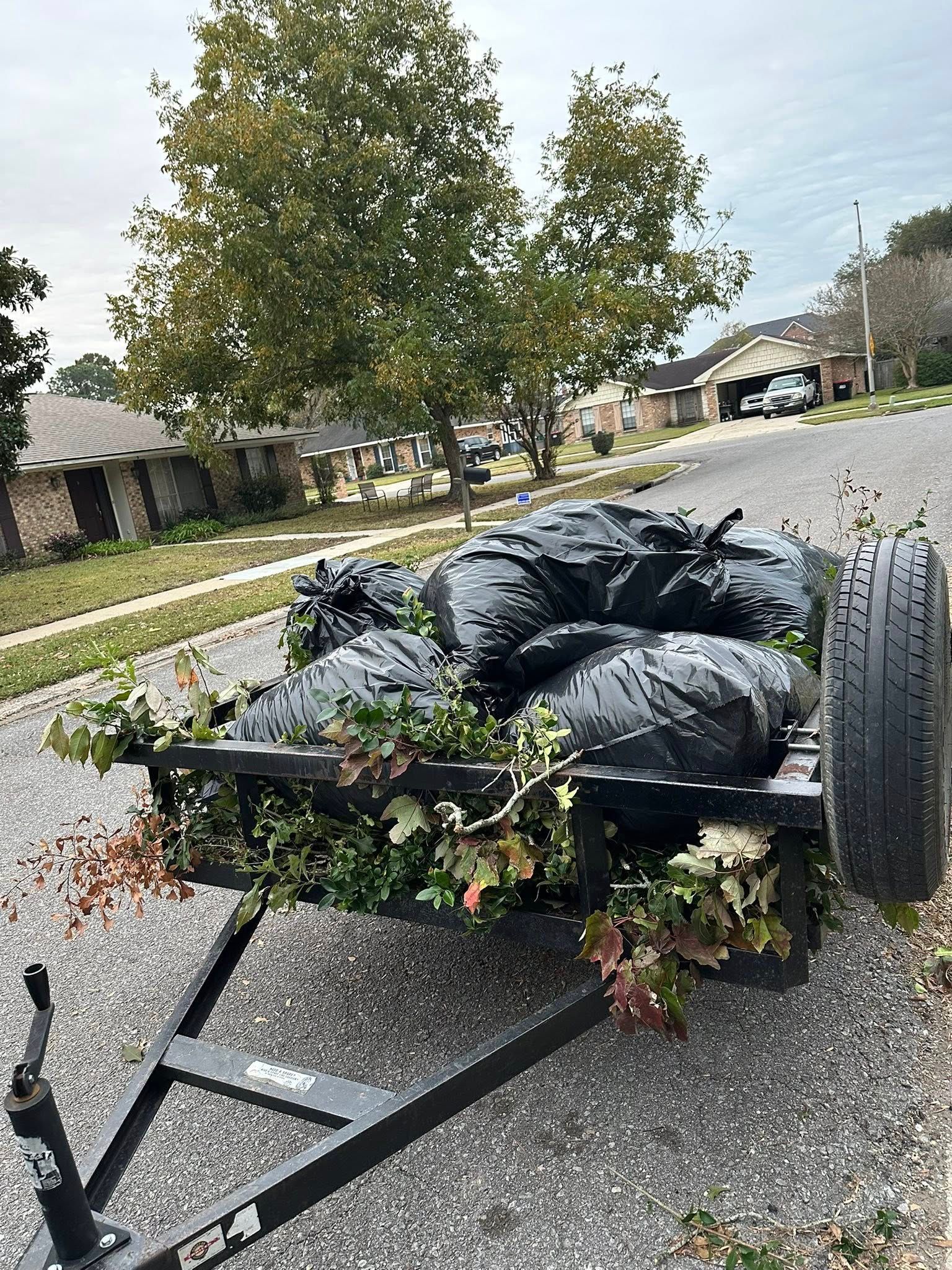 A black utility trailer loaded with bags of yard waste and tree trimmings parked on a residential street.